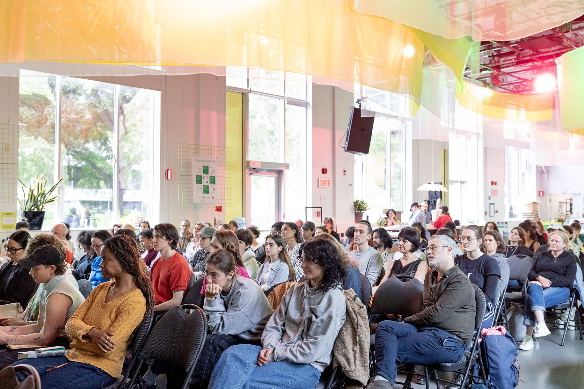 Une assemblée de personnes est assise sur des rangés de chaises et écoute un conférencier qu'on ne voit pas à l'image.
