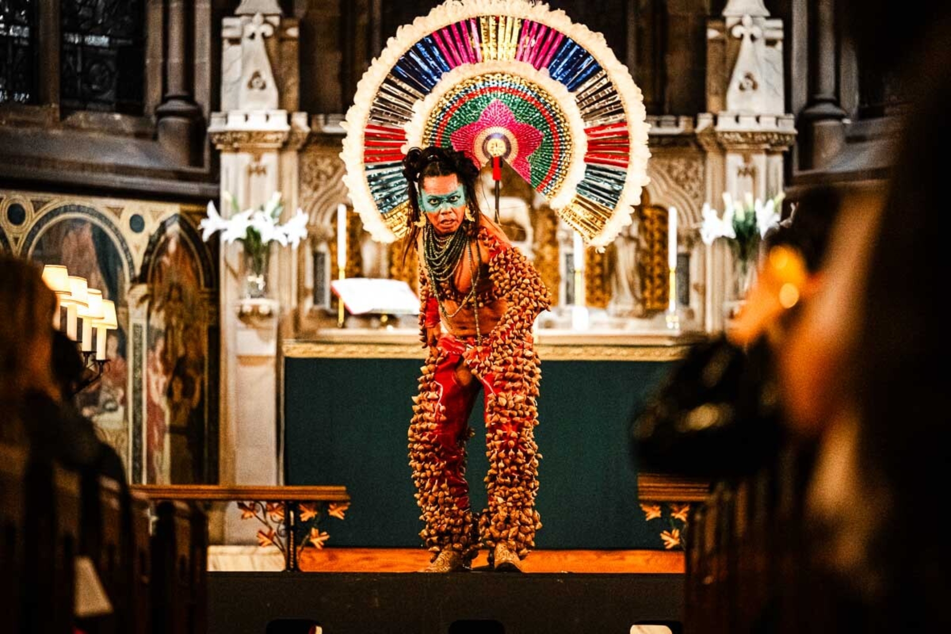 Lukas Avendaño est habillé de manière très colorée avec un maquillage bleu sur les yeux et s'avance vers le public dans une chapelle.