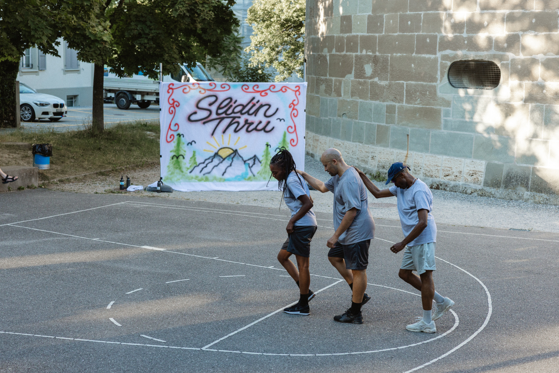 3 danseurs en t-shirts et shorts gris sur un terrain de basket appuient leur main droite sur l'épaule droite de la personne devant elleux. Au fond, on voit un tissu blanc tendu où il est inscrit Slidin Thru comme sur un tag.