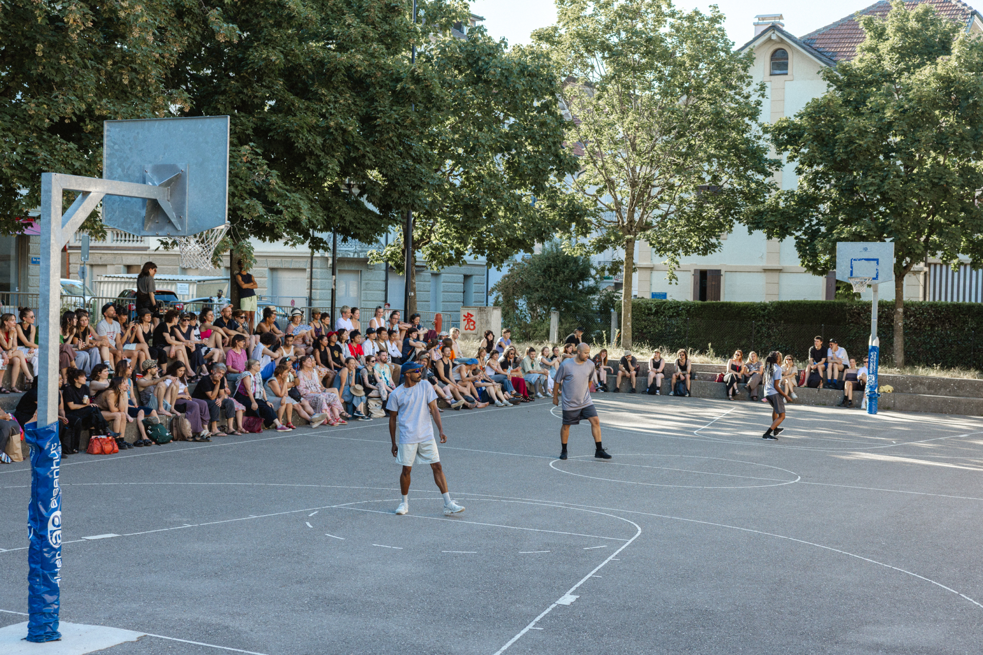 3 danseurs en shorts et t-shirts gris sont sur un terrain de basketball et un public est rassemblé en bordure du terrain.