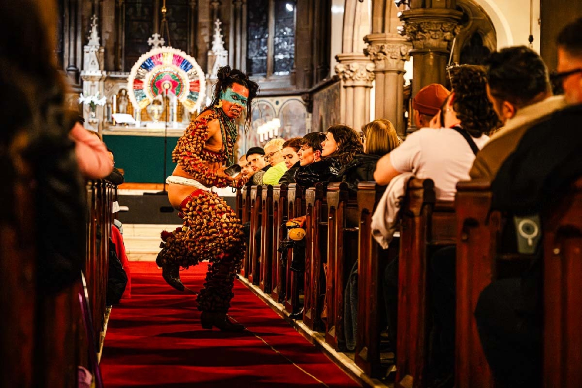 Lukas Avendaño est habillé de manière très colorée avec un maquillage bleu sur les yeux et s'avance en dansant vers le public dans une chapelle.