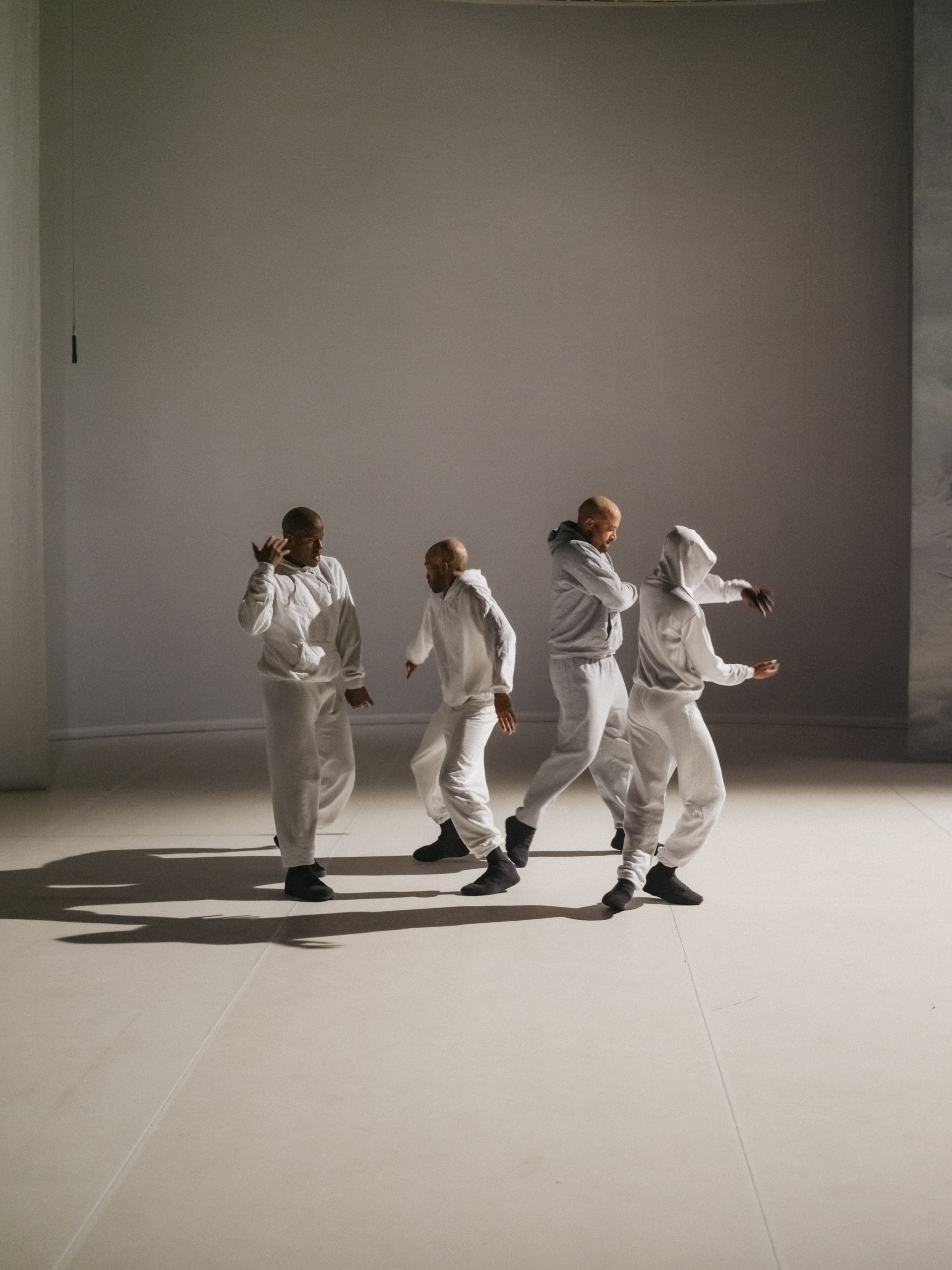 4 danseurs en ensemble de jogging blanc sont regroupés sur une scène blanche éclairé depuis la droite par une lumière ce qui crée un jeu d'ombres.