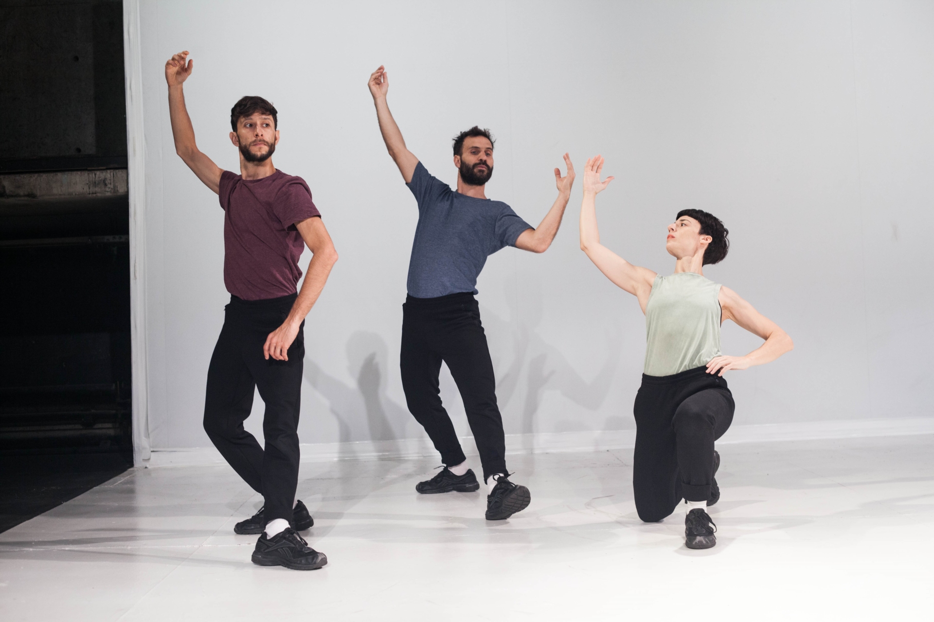 Trois danseurs en pantalon noir et t-shirt pastel lèvent leurs bras vers le haut en reculant leur buste au milieu d'une scène blanche et vide.
