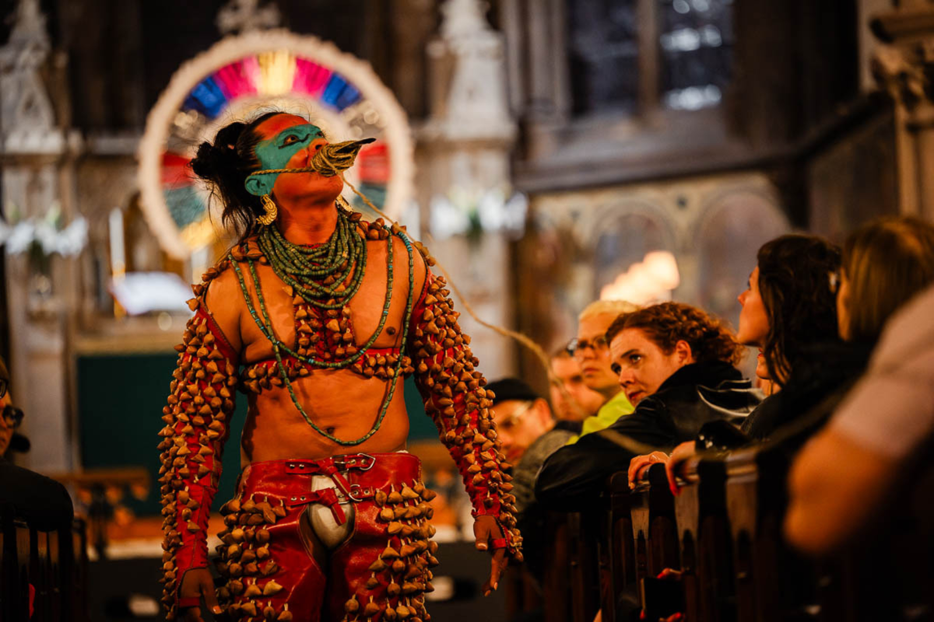 Lukas Avendaño est habillé de manière très colorée avec un maquillage bleu sur les yeux et incline sa tête vers le toit d'une chappelle, au milieu du corridor entre les rangées de spectateurs.
