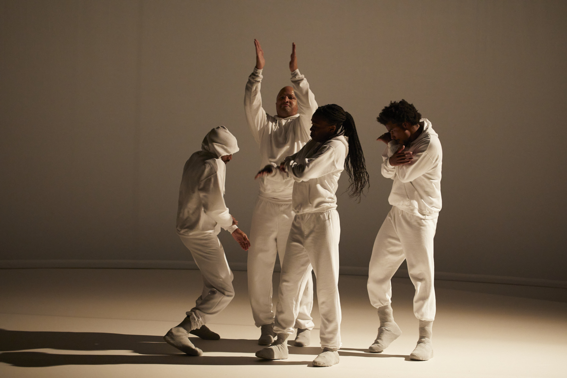 4 danseurs en ensemble de jogging blanc sont regroupés sur une scène blanche éclairé depuis la droite par une lumière chaude ce qui crée un jeu d'ombres.