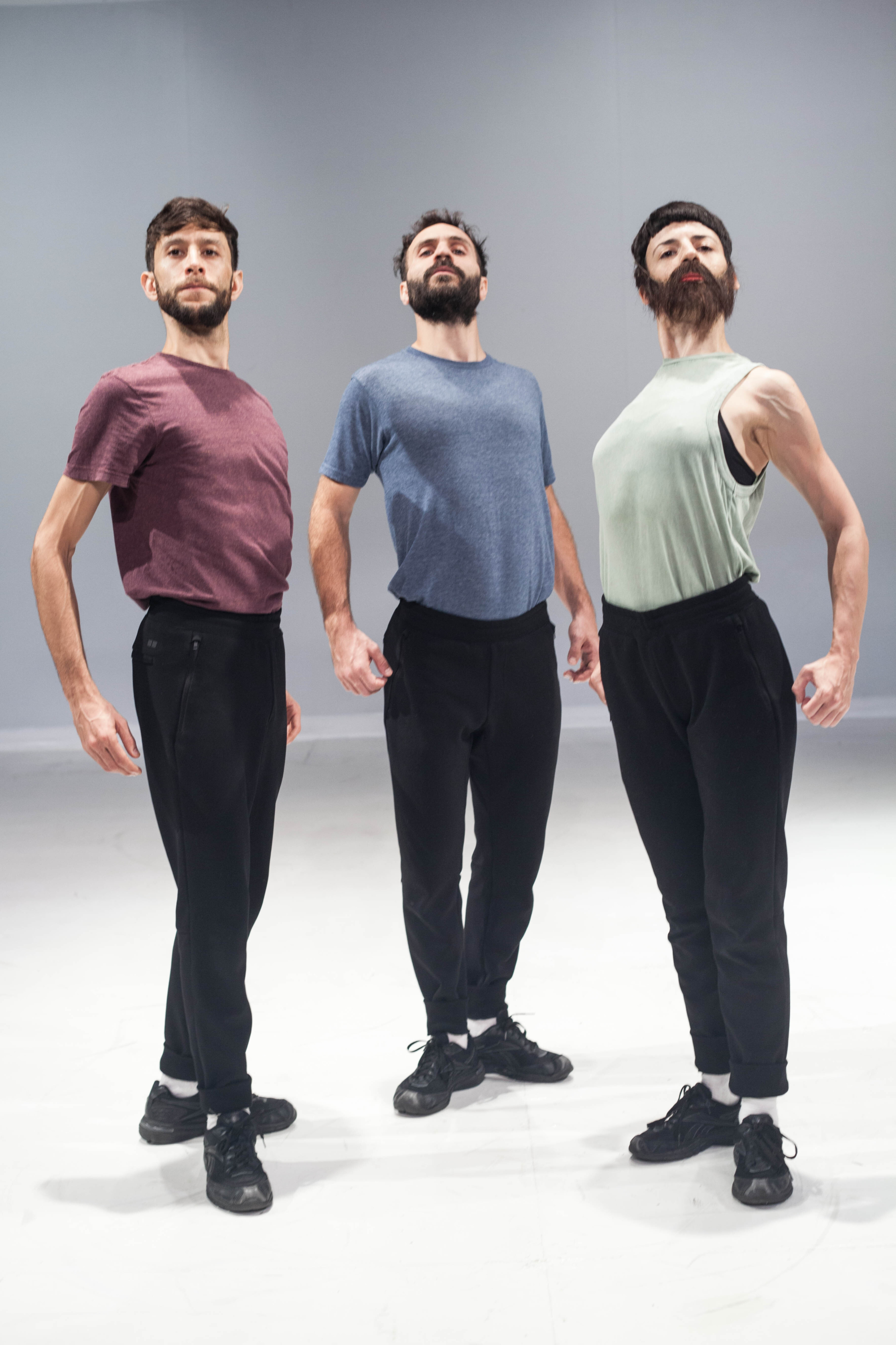 Trois danseurs en pantalon noir et t-shirt pastel bombent le torse au milieu d'une scène blanche et vide.