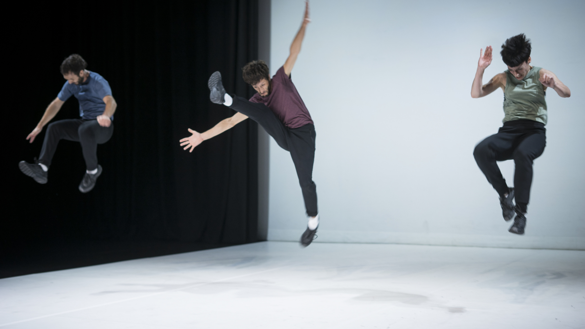 Trois danseurs en pantalon noir et t-shirt pastel sautent en l'air dans des positions différentes.