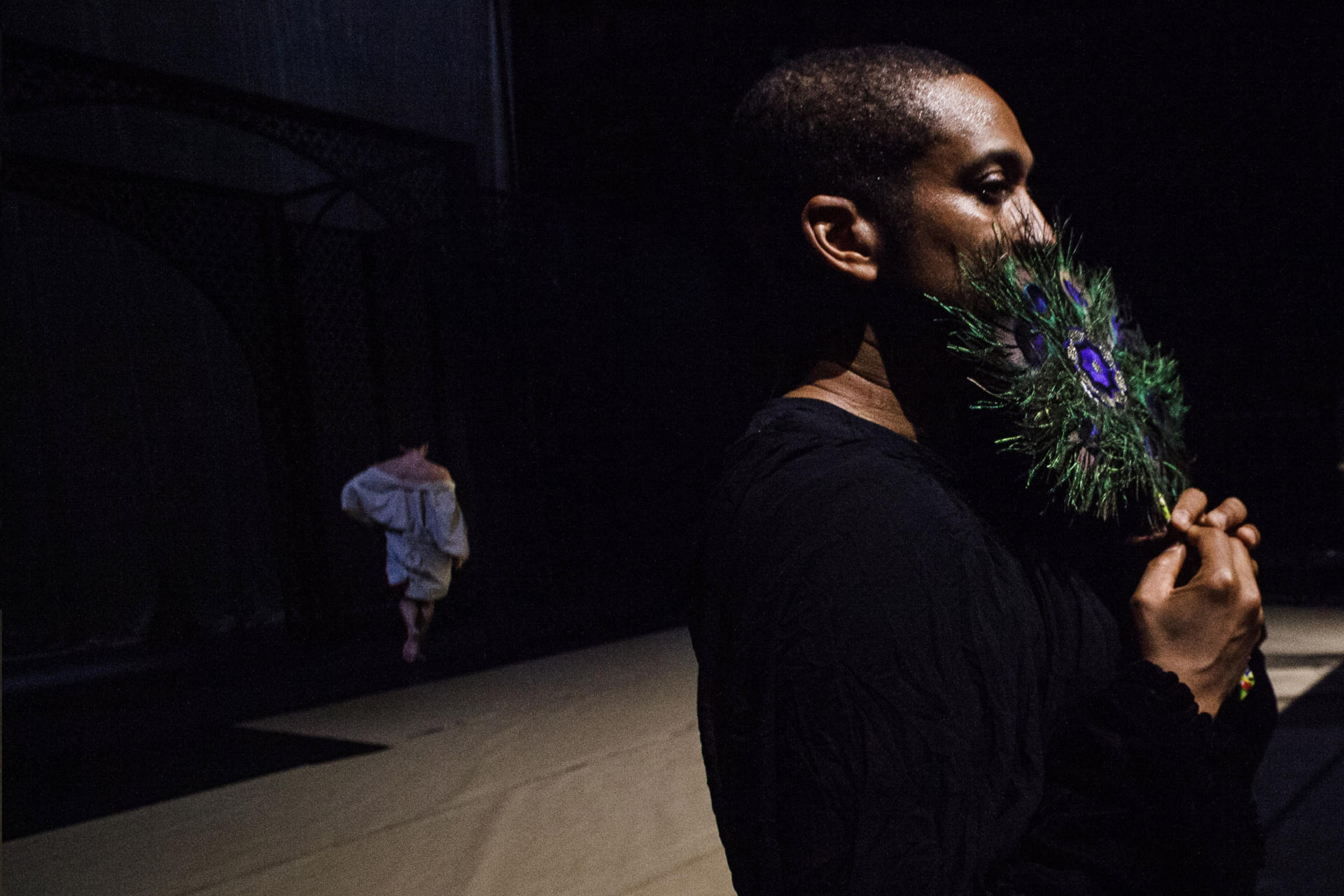 Choreographer Trajal Harrell is shown in profile, dressed in black, covering the lower part of his face with the tip of a peacock feather.