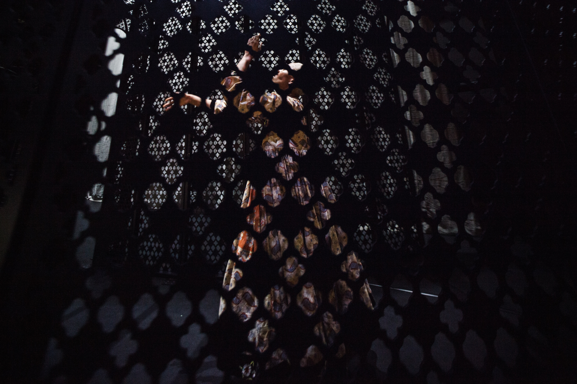 An interpreter dances behind a mashrabiya, wearing a long, patterned, pleated dress.