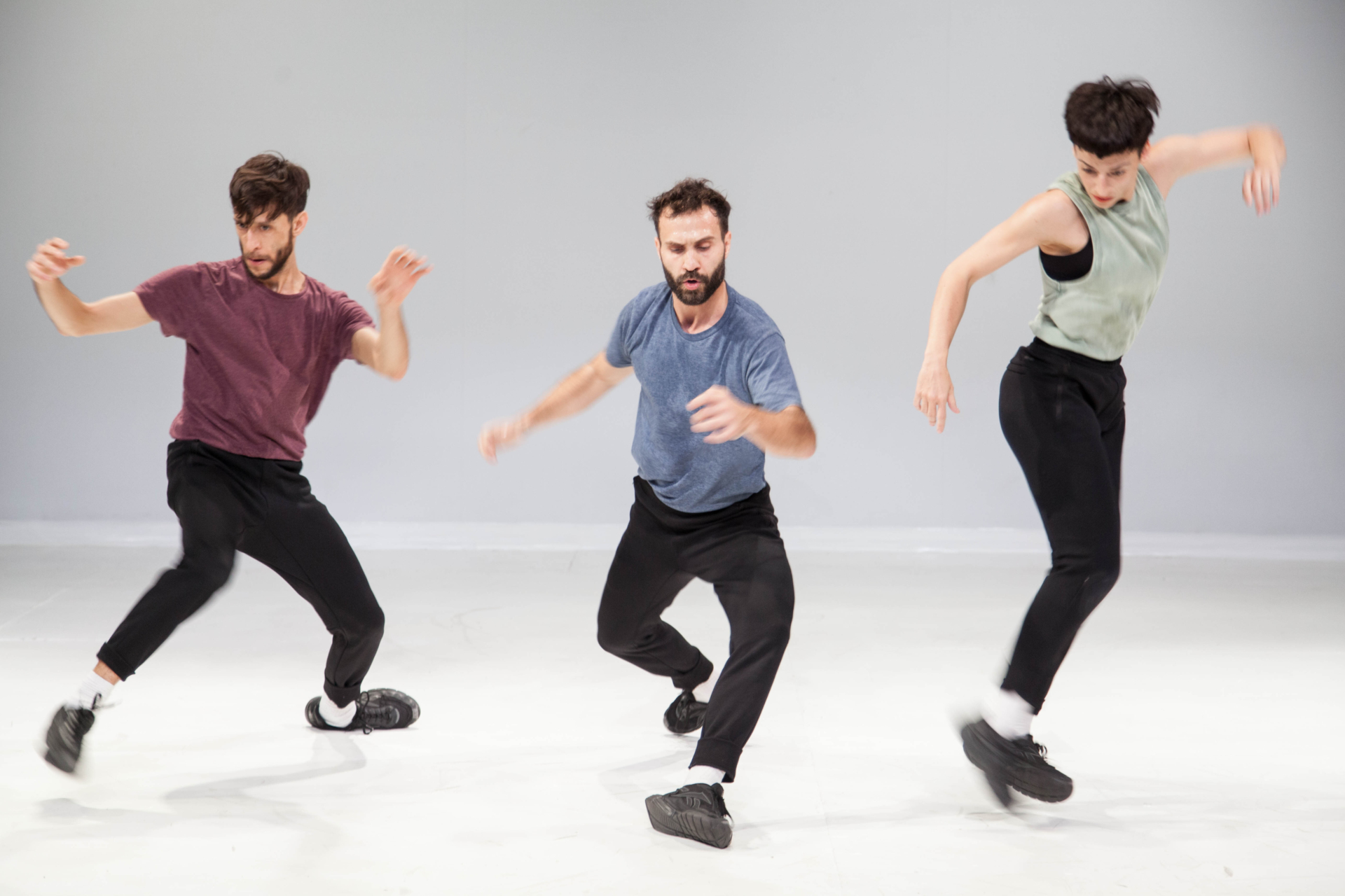 Trois danseurs en pantalon noir et t-shirt pastel sont en mouvement de rebond au milieu d'une scène blanche et vide.