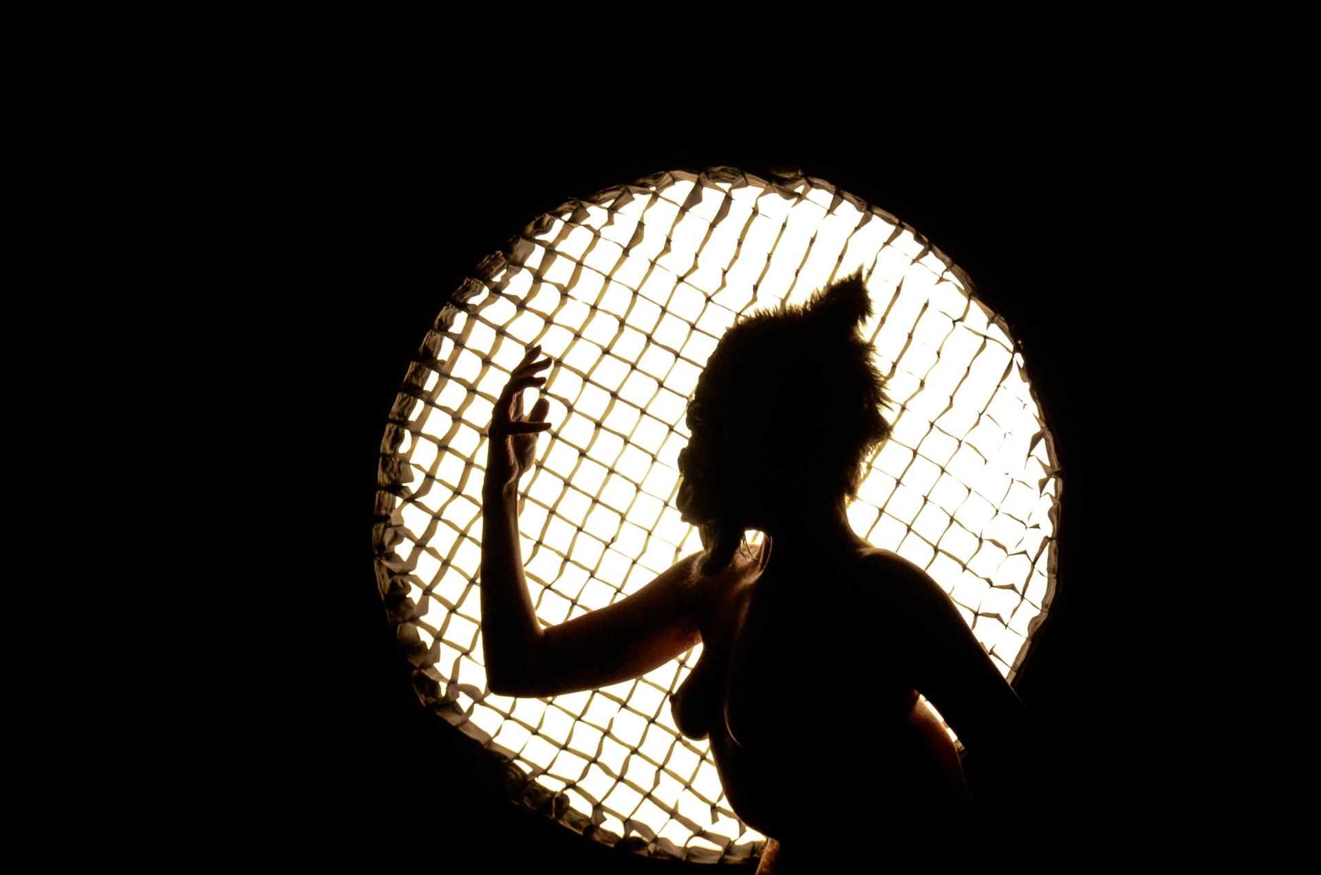 Une silhouette féminine et masquée lève son bras droit devant un cercle de lumière blanche qui la rend à contre jour.