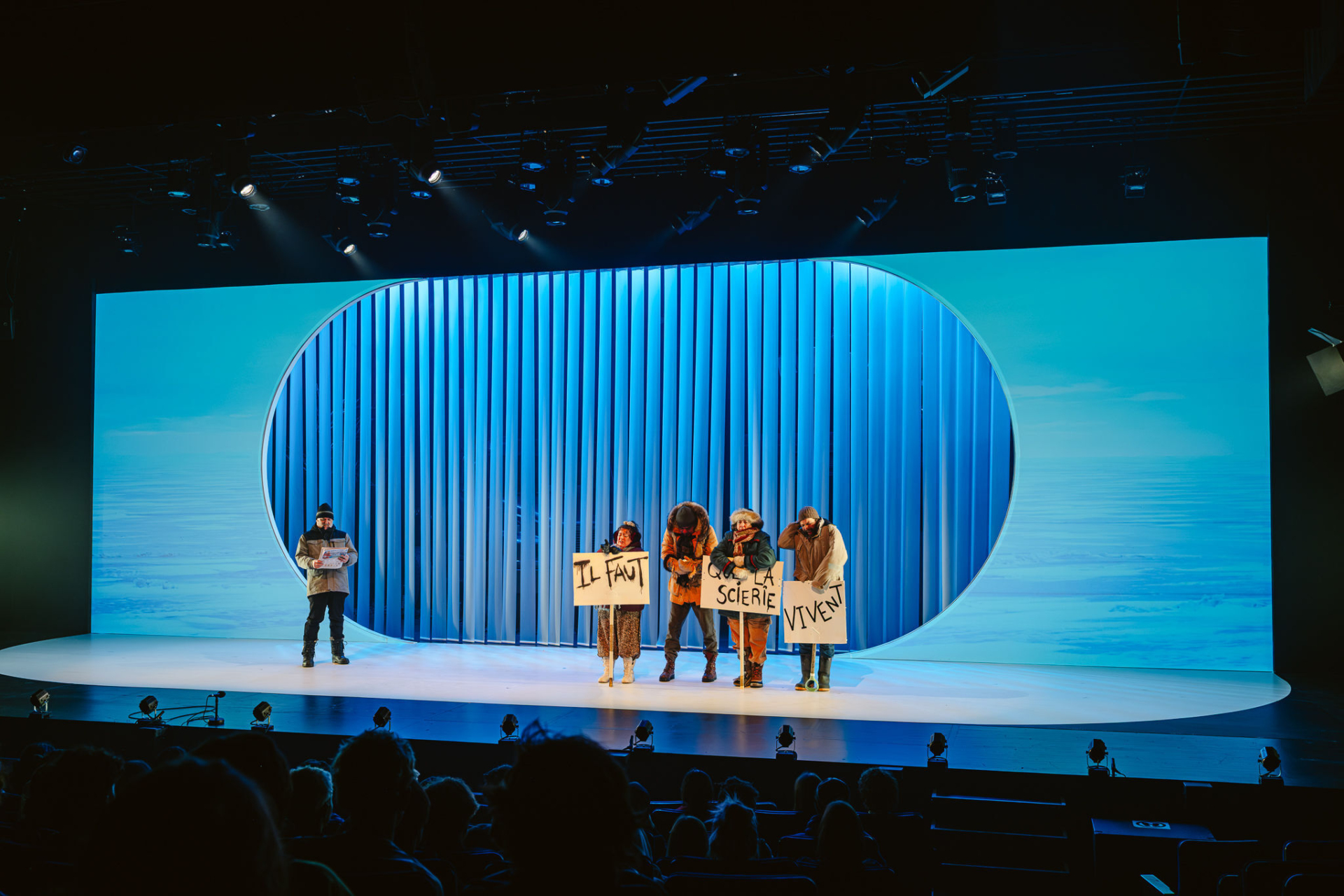 Several people holding protest signs are on a large stage with a blue-lit backdrop.