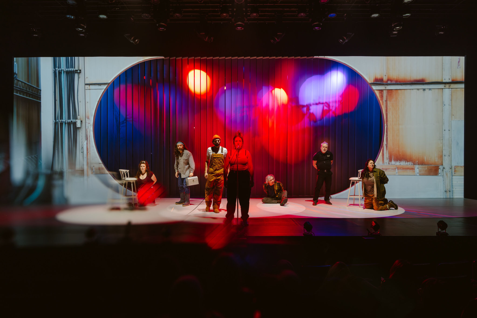 Several characters from the novel Querelle de Roberval are on stage against a backdrop resembling a hangar, lit in red.
