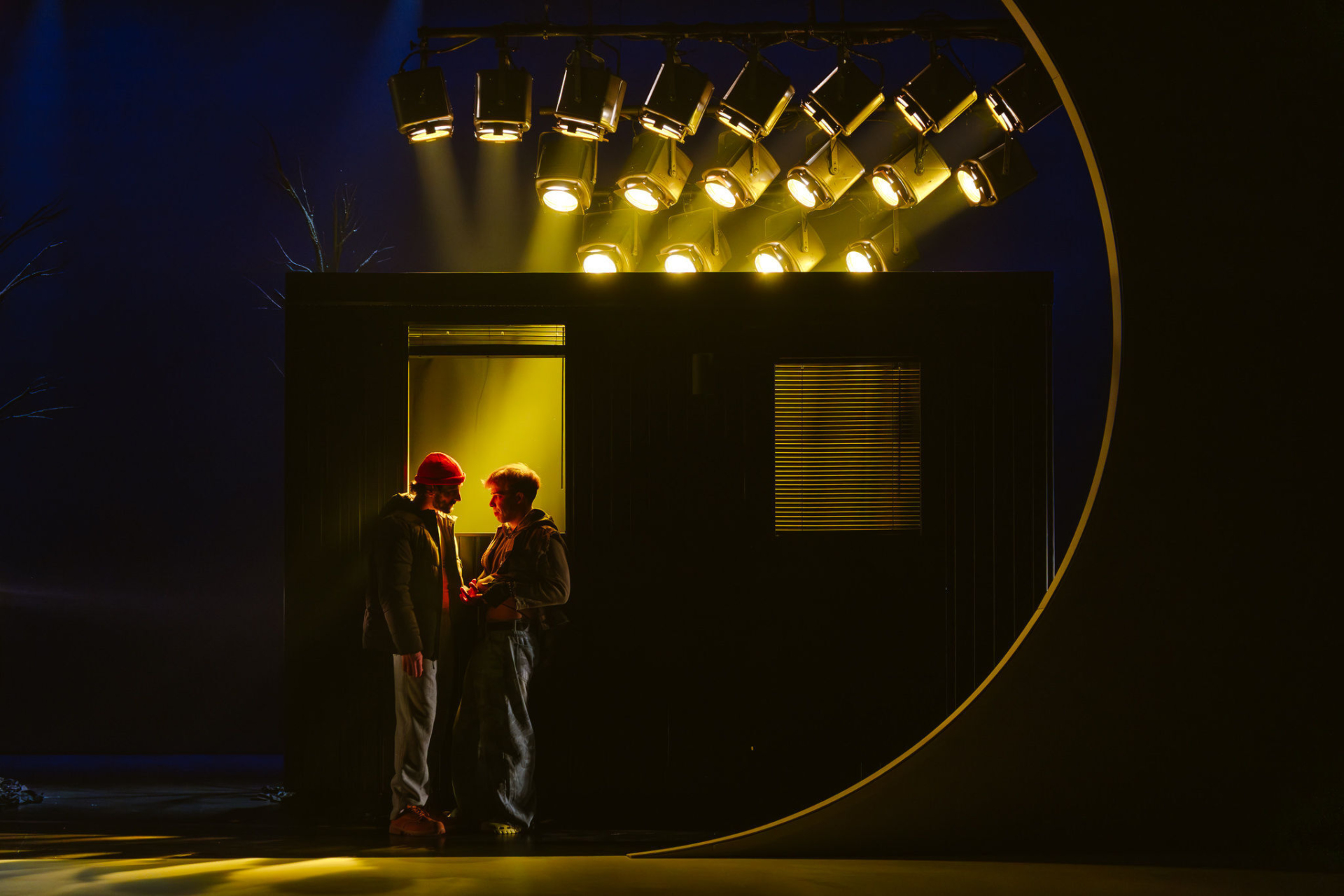 On stage, two people are leaning in the darkness against a window lit by yellow spotlights.