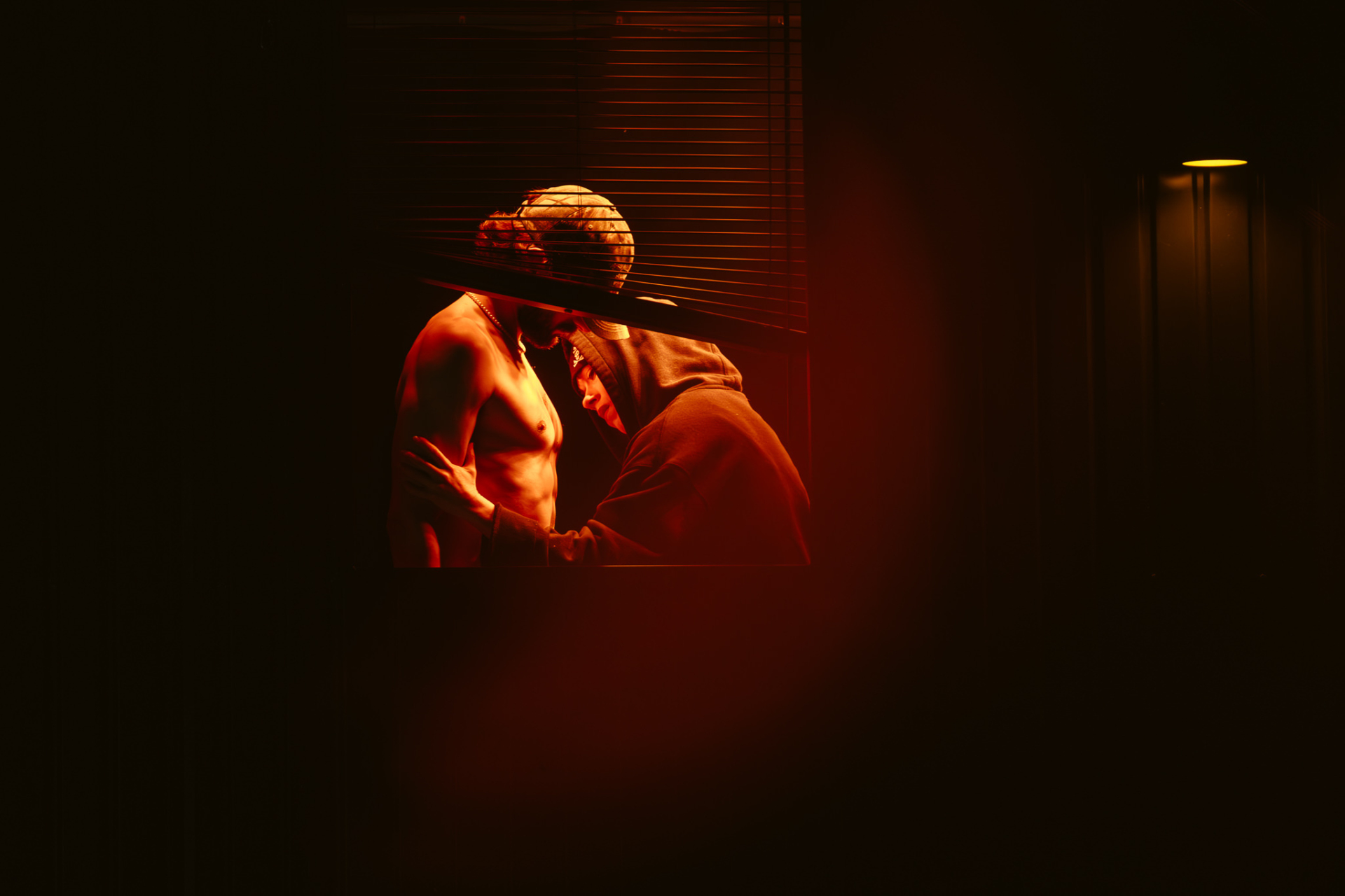 The character Querelle is seen behind the blinds of a window, lit by a warm light and shirtless, while another character approaches his torso.