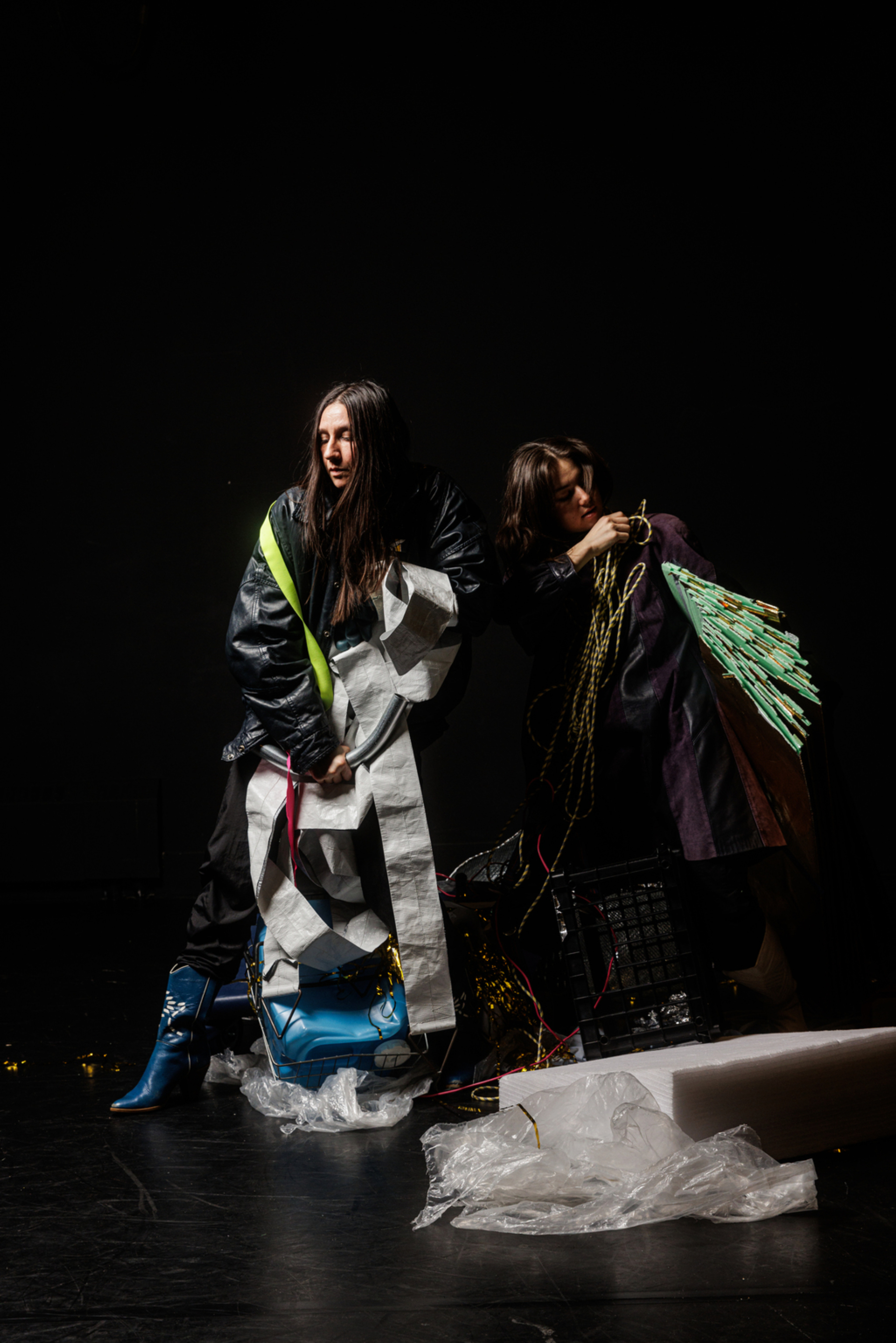 Deux performeurs vêtues de noir sur scène se couvrent d'objets en tout genre.