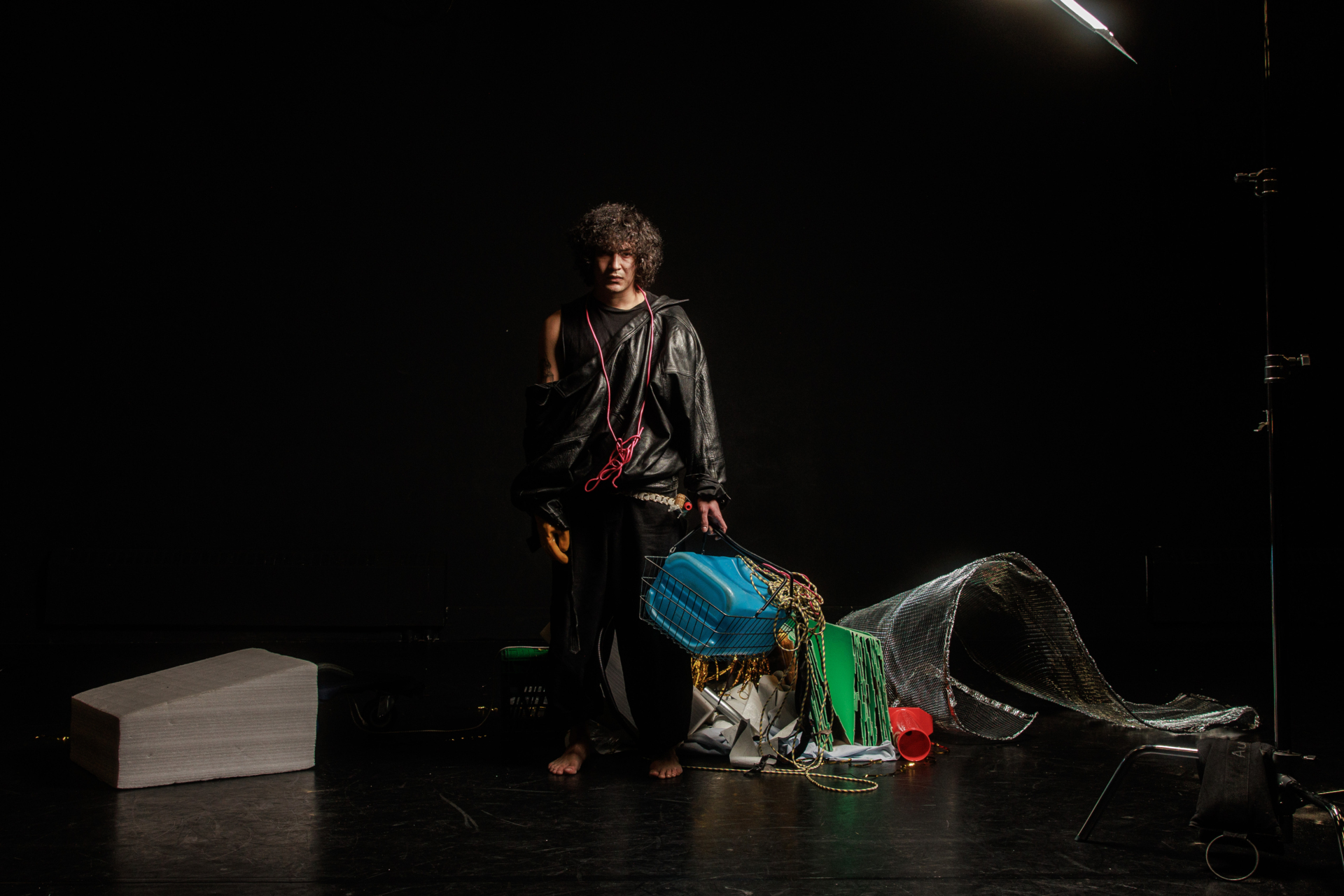 Un personnage vêtu de noir sur scène est au milieu d'un débarras d'objets et tient un panier qui contient un bidon bleu turquoise.