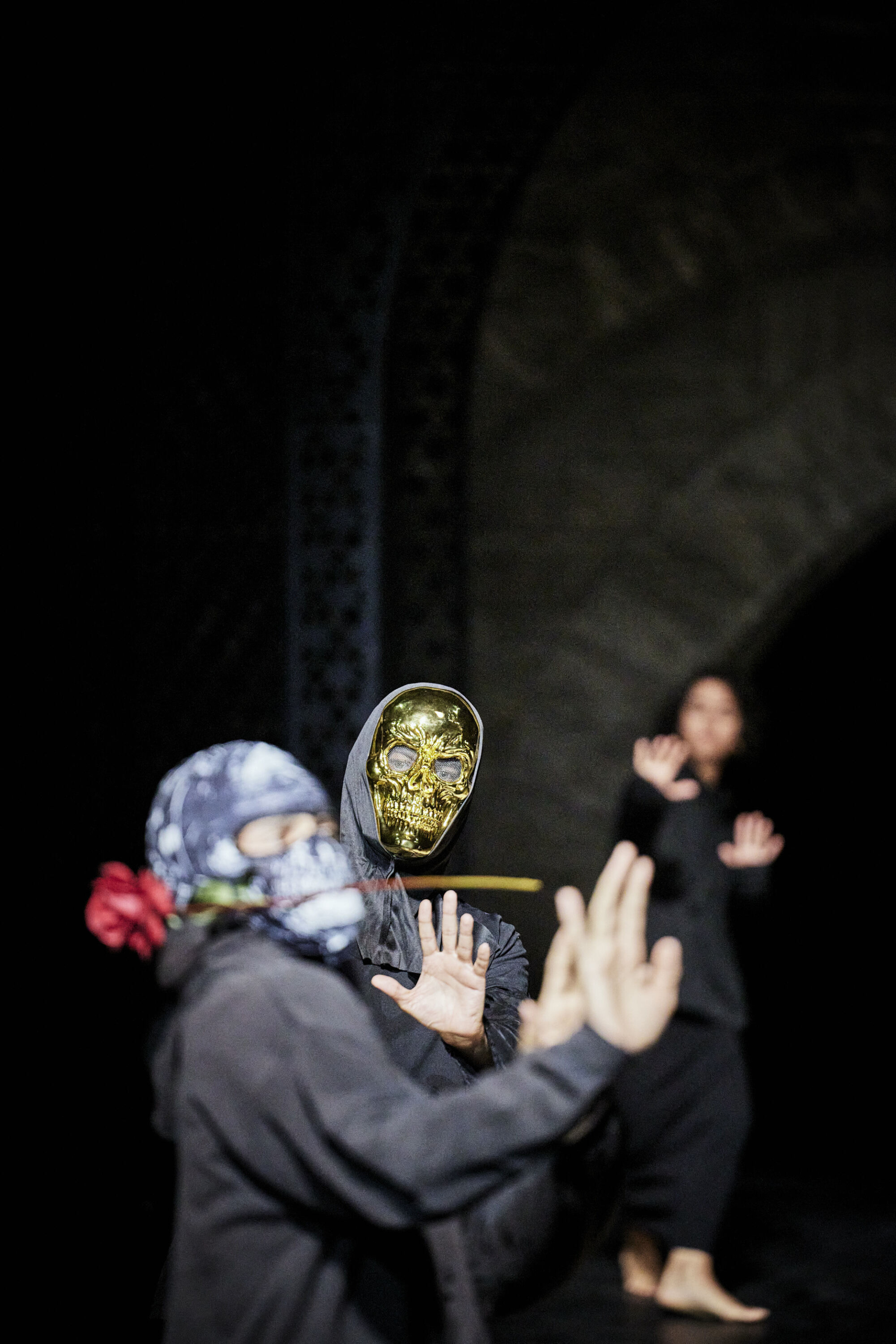 Two performers are wearing masks: the one in the foreground is wearing a hood and holding a red rose in his mouth, while the one in the background is wearing a golden skeleton mask.