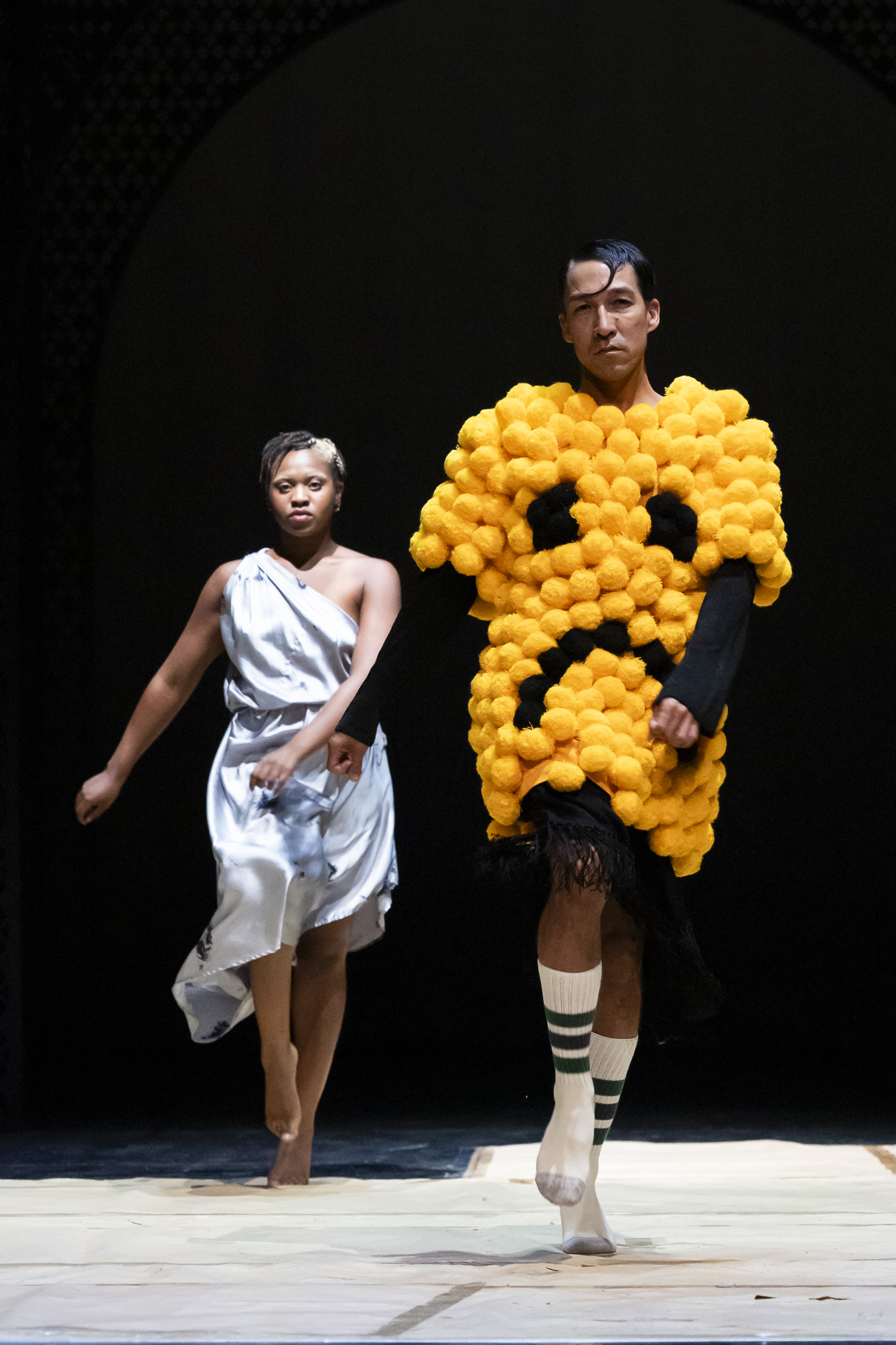 Two dancers walk toward us as if in a fashion show, and the first one is wearing a large T-shirt made of yellow balls that form a non-smiling face.