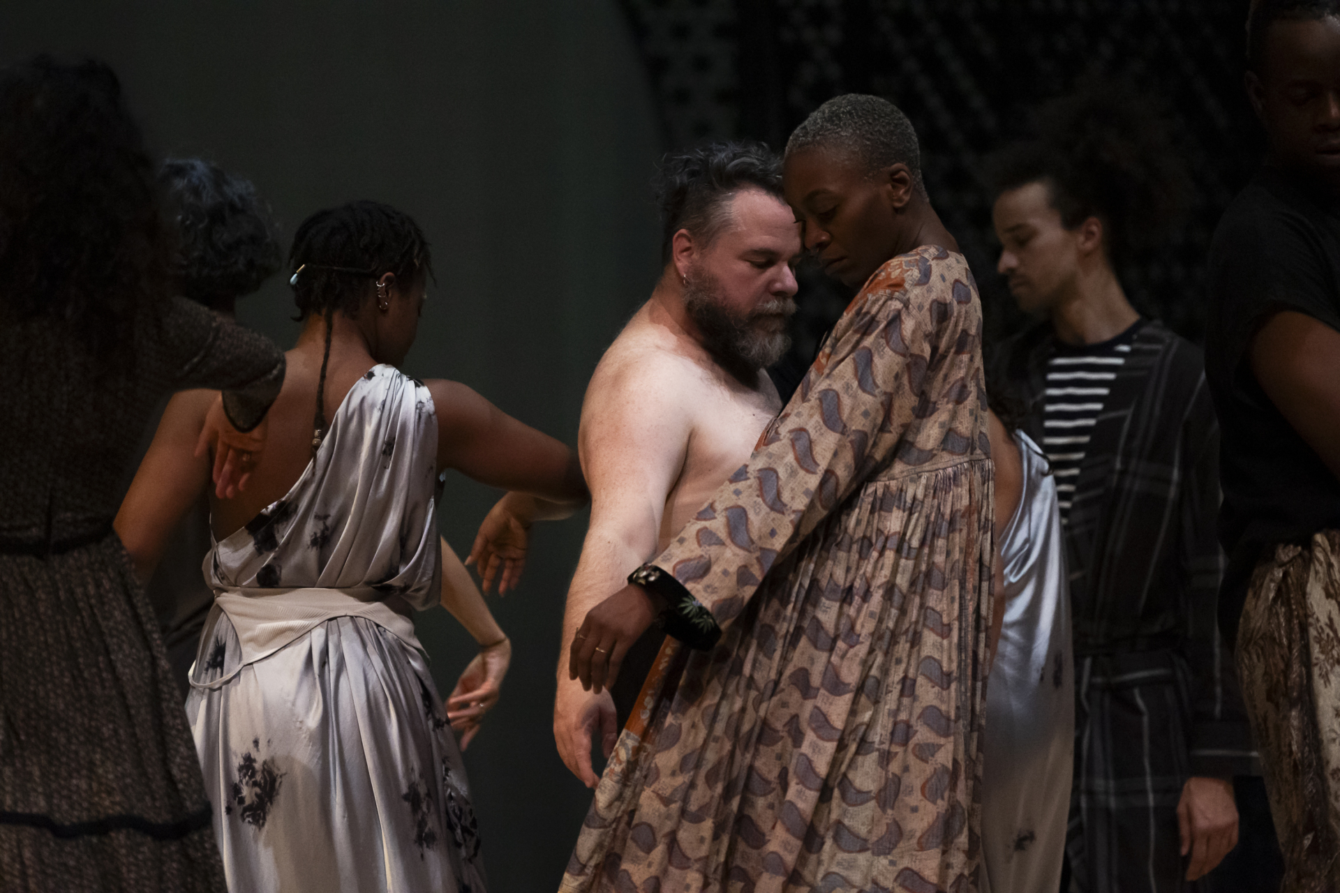 Several performers are grouped together on stage, brushing against each other, dressed in long, pleated tunics or bare-chested.