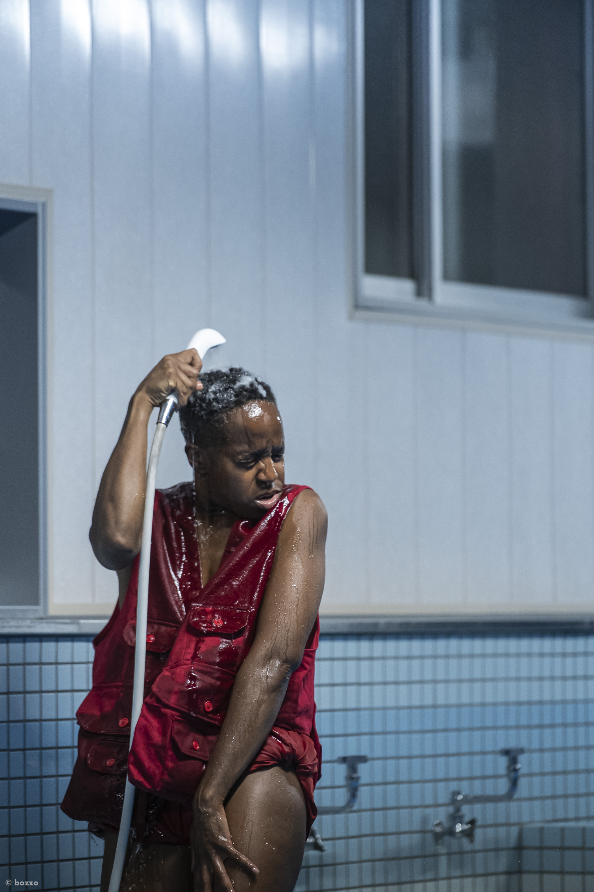 L'artiste Dana Michel vêtue d'un gilet rouge s'arrose la tête avec un pommeau de douche.