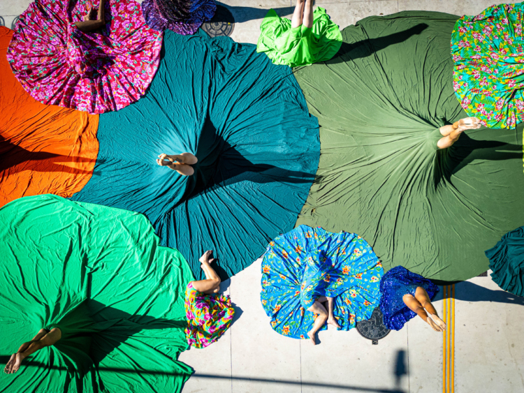 Une vue aérienne montre des cercles de tissus colorés étalés au sol. Au centre de chaque cercle se trouve une pair de jambe pointée vers le ciel.
