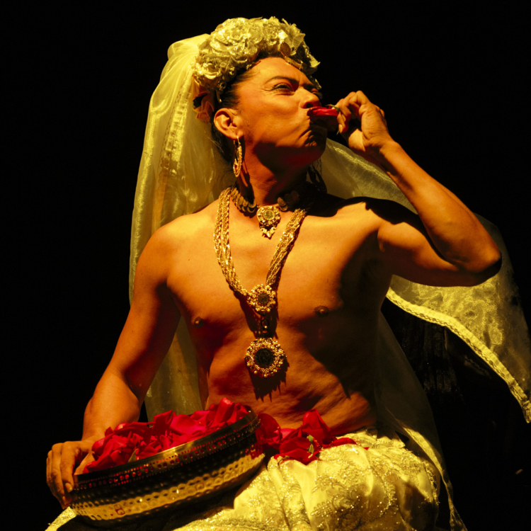 Lukas Avendaño porte une traîne blanche, un sautoir doré sur son torse nu et un récipient métallique plat alors qu'il porte une objet rouge à sa bouche, pratiquant une sorte de rituel ancestral.