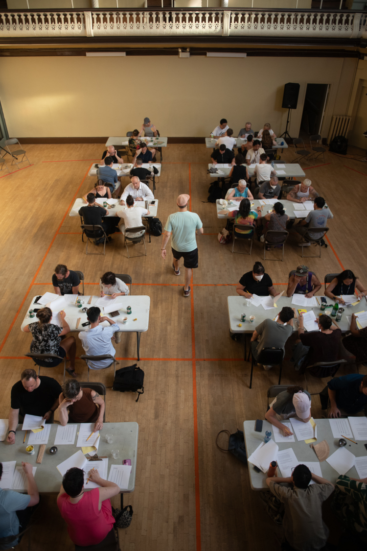 Plusieurs personnes sont assises par groupes de 4 autour de tables dans une grande salle et un acteur circule entre elles.