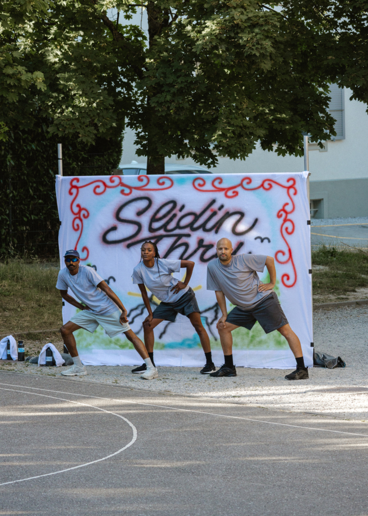 3 danseurs en t-shirts et shorts gris au bord d'un terrain de basket appuient leur main droite sur leur genou droit devant un tissu blanc tendu où il est inscrit Slidin Thru comme sur un tag.