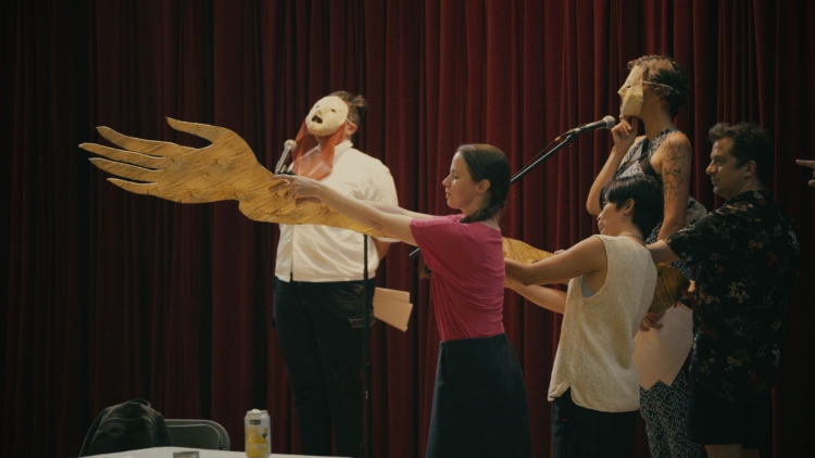 Des participants tiennent une grande main en or alors que des personnes avec des masques en papier parlent.