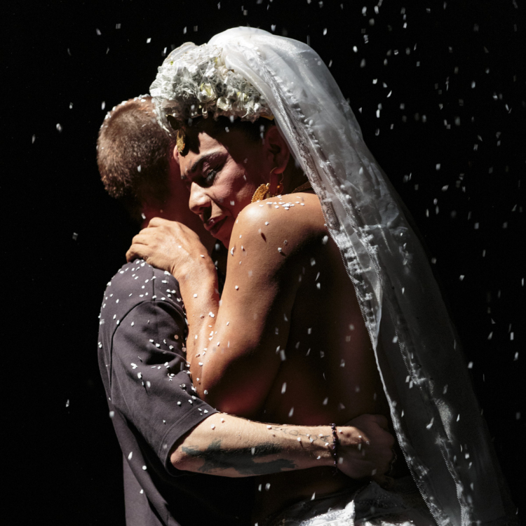 Lukas Avendaño porte une traîne blanche et enlace un membre du public alors que des flocons de papier tombent sur eux.