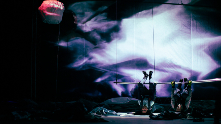 Deux personnages en costume sont suspendus à une barre métallique sur une scène éclairée en noir et violet