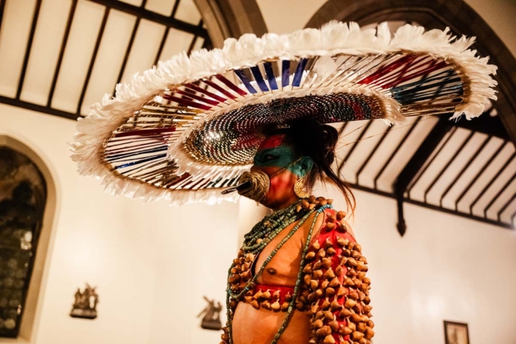 Lukas Avendaño est habillé de manière très colorée avec un maquillage bleu sur les yeux et porte une sorte de grande coiffe sur le dessus de son crâne.