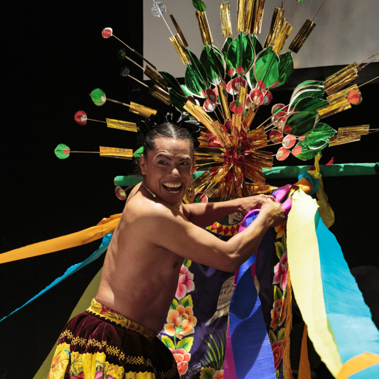 Lukas Avendaño est torse nu, avec une jupe colorée et se tient devant une sculpture métallique multicolore a laquelle il accroche de grands rubans.
