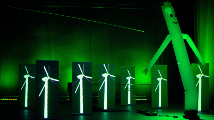 Sur une scène éclairée par une lumière verte, des images d'éoliennes sont projetées sur une multitude d'écran en tissu verticaux accompagné d'un bonhomme gonflable publicitaire