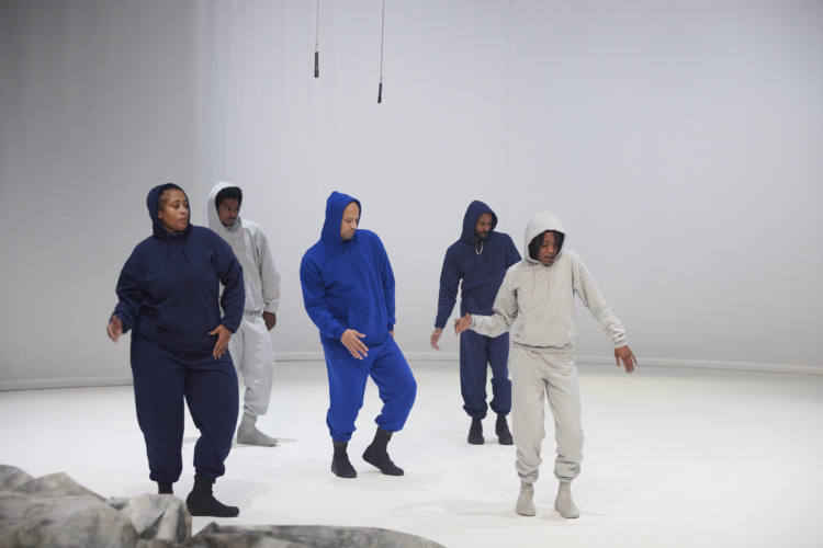 Cinq danseurs et danseuses en ensemble jogging gris, noir ou bleu sont sur une scène très blanche avec deux micros suspendus au plafond.
