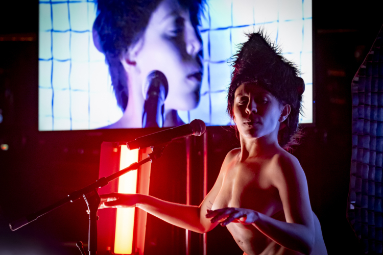 Une personne dont on voit la poitrine nue se tient devant un micro avec une sorte de masque poilu qui recouvre ses cheveux.