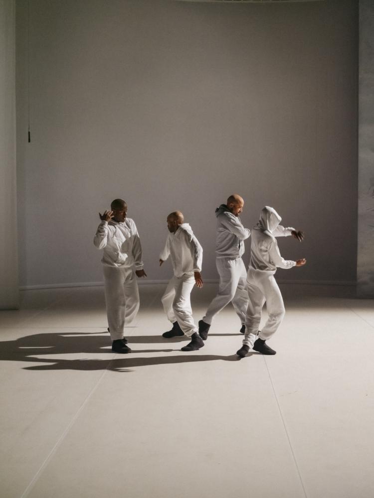 4 danseurs en ensemble de jogging blanc sont regroupés sur une scène blanche éclairé depuis la droite par une lumière ce qui crée un jeu d'ombres.