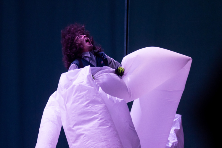 A character with long, curly hair emerges from an inflatable advertising figure illuminated by purple light, shouting