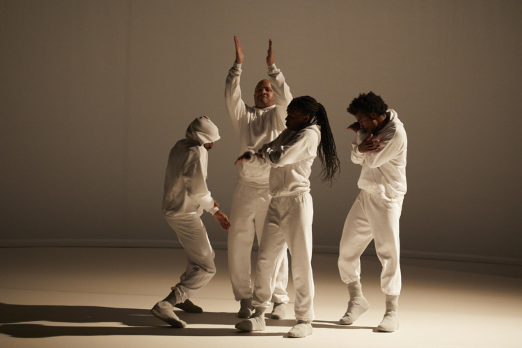 4 danseurs en ensemble de jogging blanc sont regroupés sur une scène blanche éclairé depuis la droite par une lumière chaude ce qui crée un jeu d'ombres.