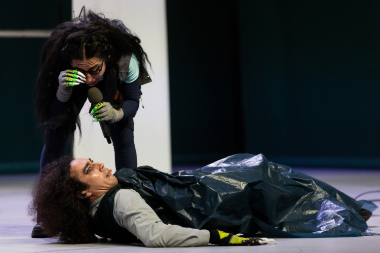 Un personnage avec des griffes aux doigts et des lunettes, tenant un micro, se penchent au dessus d'un deuxième personnage allongé au sol, vêtu d'un grand sac poubelle noir