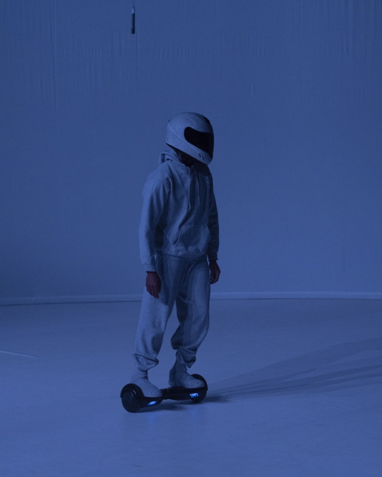 Une personne sur un hoverboard, vêtue d'un survêtement gris clair et d'un casque, se déplace dans un espace entièrement bleu.