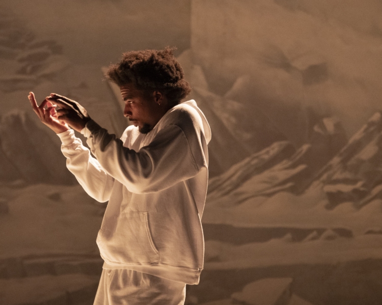 Un danseur vêtu d'un survêtement blanc évolue devant un décor représentant des montagnes, les bras levés.