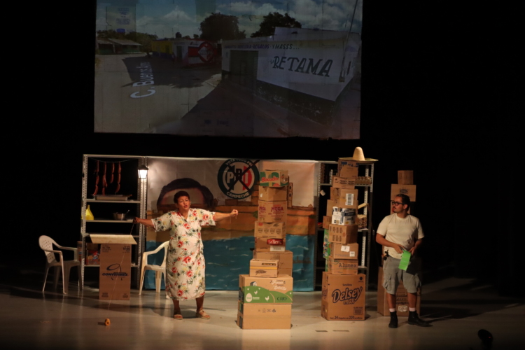 On a stage with stacks of cardboard boxes, Josefina Orlaineta stretches out her arms while her son watches her