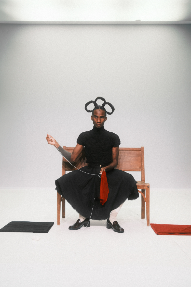 Assis sur un banc, Mackenzy Bergile, vétu d'un col roulé et d'une jupe noir·e, coud en regardant directement la caméra.