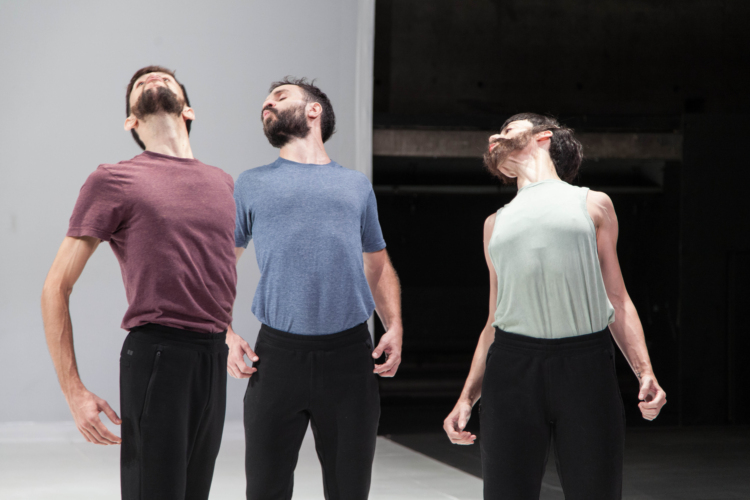 Trois danseurs en pantalon noir et t-shirt pastel relèvent leur tête vers le haut en faisant ressortir leur buste, les bras arqués.