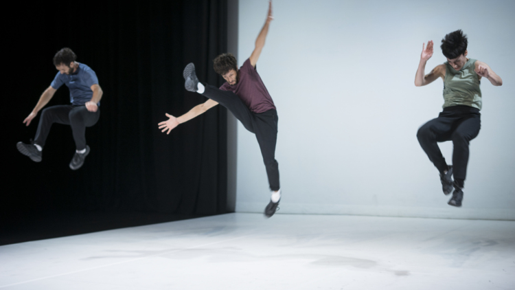Trois danseurs en pantalon noir et t-shirt pastel sautent en l'air dans des positions différentes.