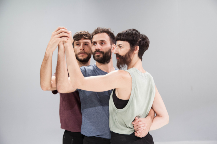 Trois danseurs en pantalon noir et t-shirt pastel se tiennent proches et son rassemblés par leurs bras réunis en un pour former un poing dirigé vers le ciel.