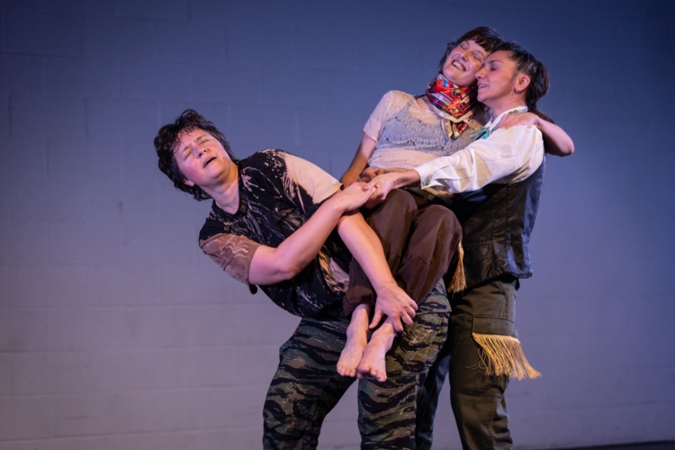 L'interprète du centre est assise sur le dos de celle de gauche alors que la personne de droite l'aide à garder l'équilibre.