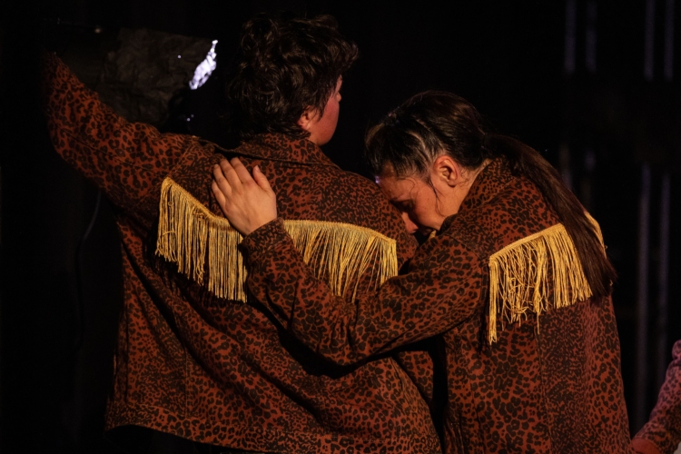 Les deux interprètes, de dos, portent des vestons bruns avec une frange jaune dans le dos. La personne de droite repose son front sur l'épaule de celle de gauche.
